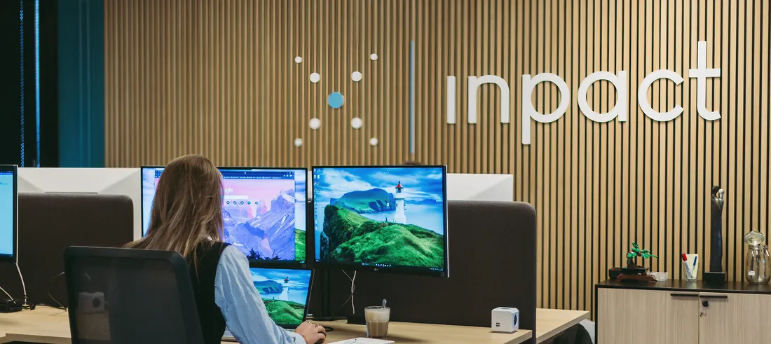 Een collega aan het werk achter drie schermen, met het Inpact-logo op de wand
